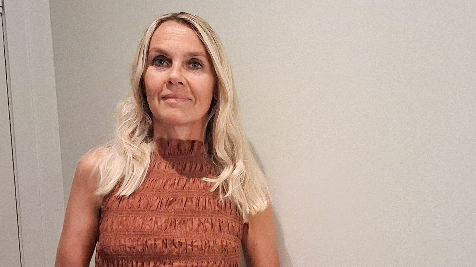 Portrettbilde av dame med blondt hår og rustfarget kjole mot hvit vegg
