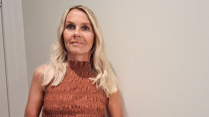 Portrettbilde av dame med blondt hår og rustfarget kjole mot hvit vegg