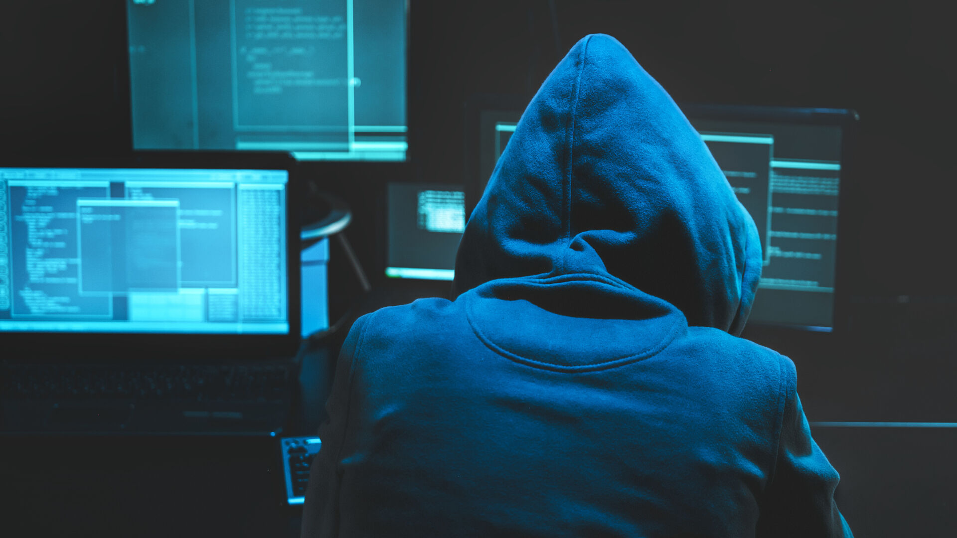 Person med hette over hodet sitter foran flere PC-skjermer og hacker