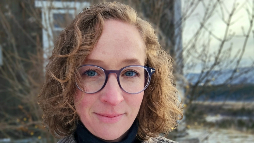 Lene ute foran et gammelt hus senhøst med briller og jakke