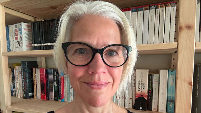 Nærbilde av eldre dame med grått hår og svarte briller mot en bokhylle
