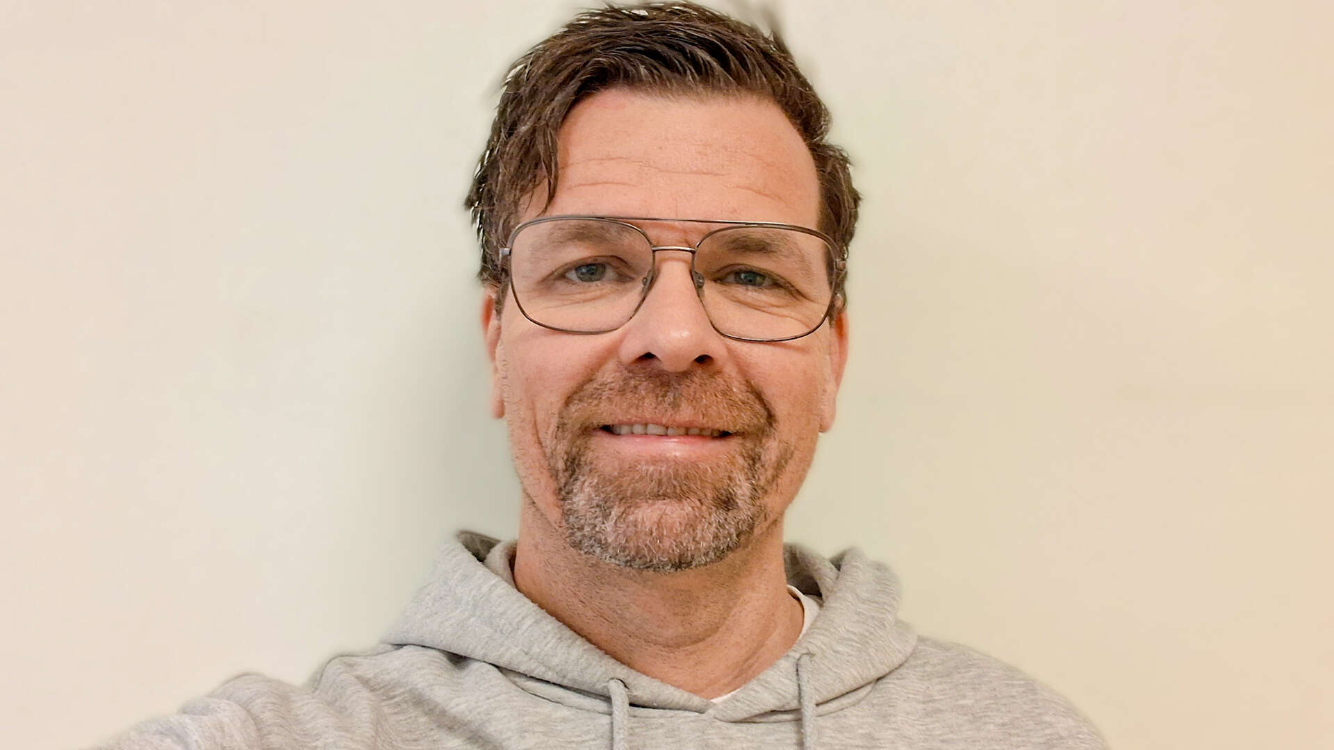 Ansiktet til en mann med briller og grå hettegenser mot en hvit vegg