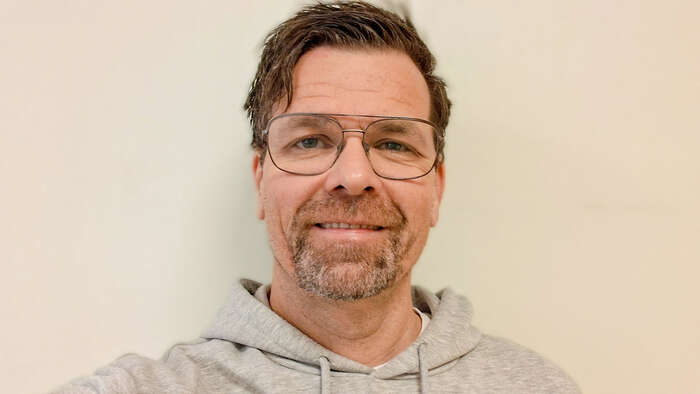 Ansiktet til en mann med briller og grå hettegenser mot en hvit vegg
