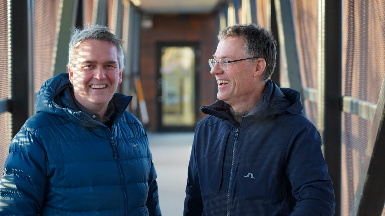 Trond og Kjell ler når de blir tattbilde av i en lang gang utendørs med gitter på hver side