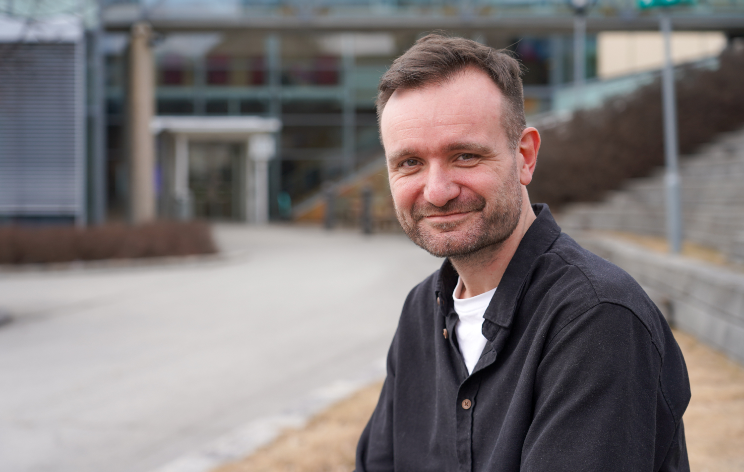 Mann sitter ute på en murkant foran et bygg med skiferplater og mange, store vinduer
