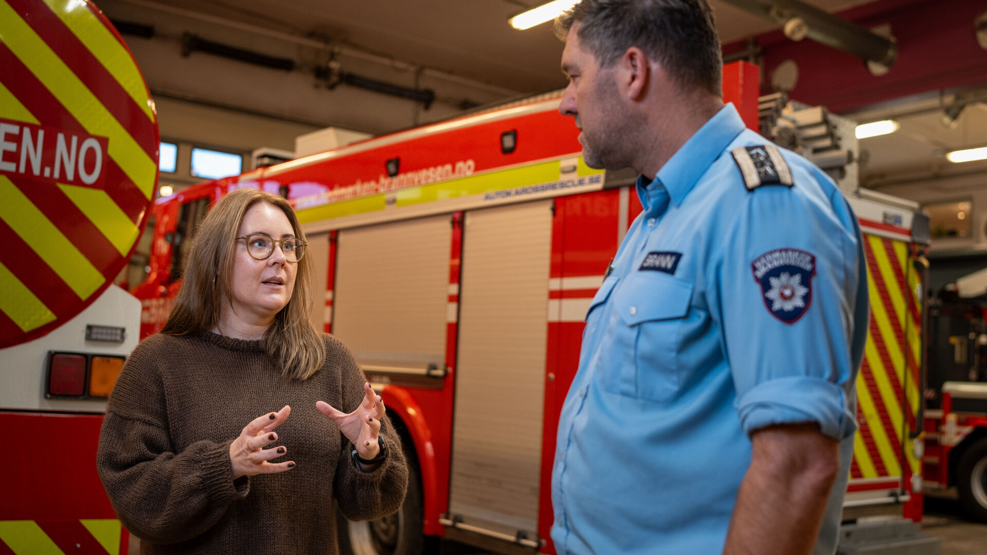 Dame med brun genser og briller snakker med en brannmann inne i en garasje med brannbiler