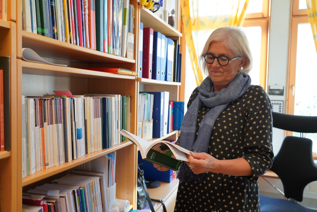 Anne Stine blar i bok inne på sitt kontor, stående ved bokhylle.