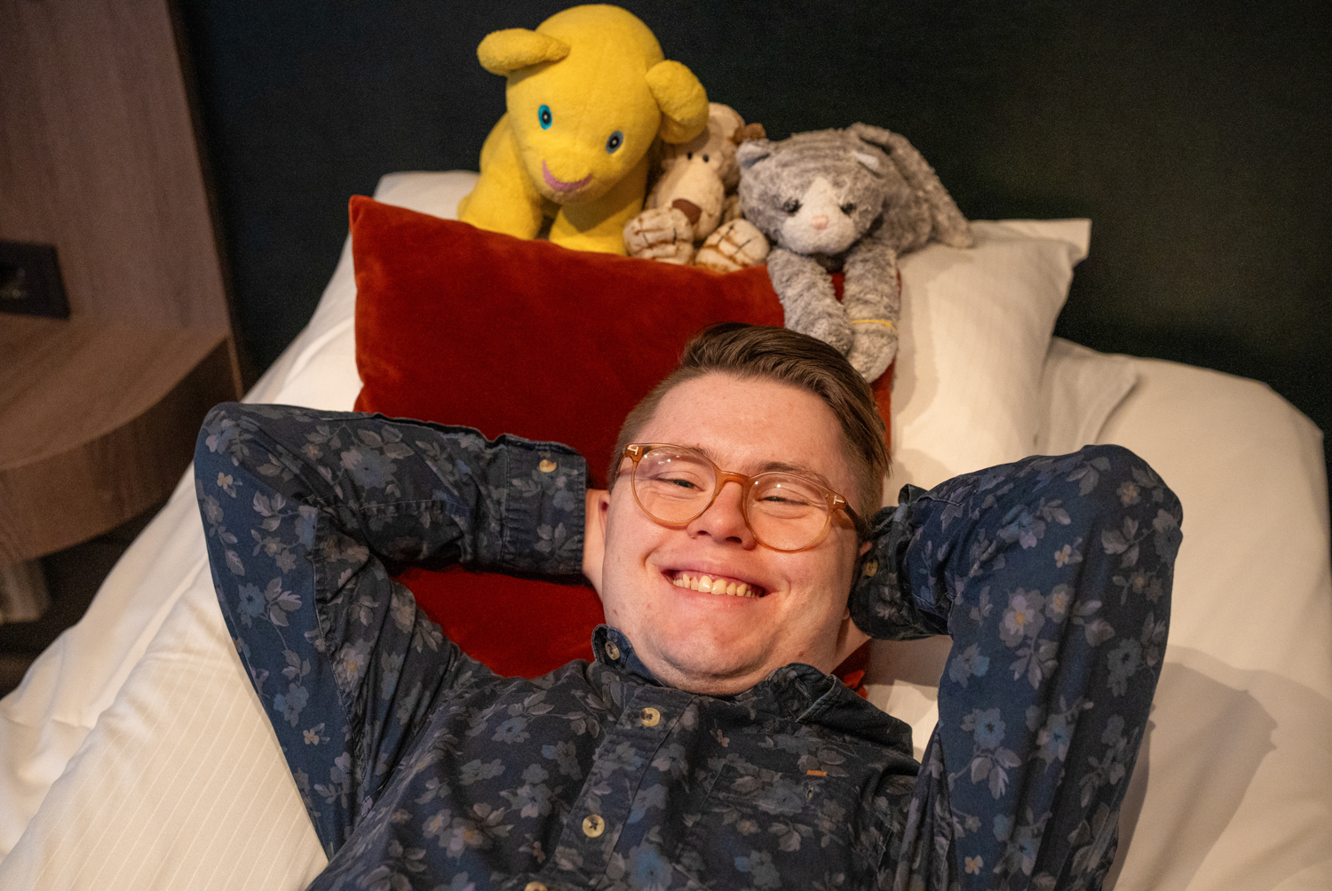 Erik ligger på sengen og smiler med kosedyrene sine bak seg.