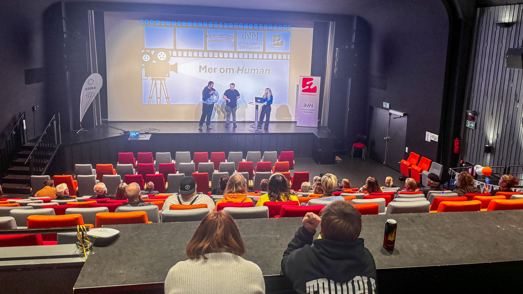 Tre personer står  på en scene på en kino med bilde på lerret bak seg