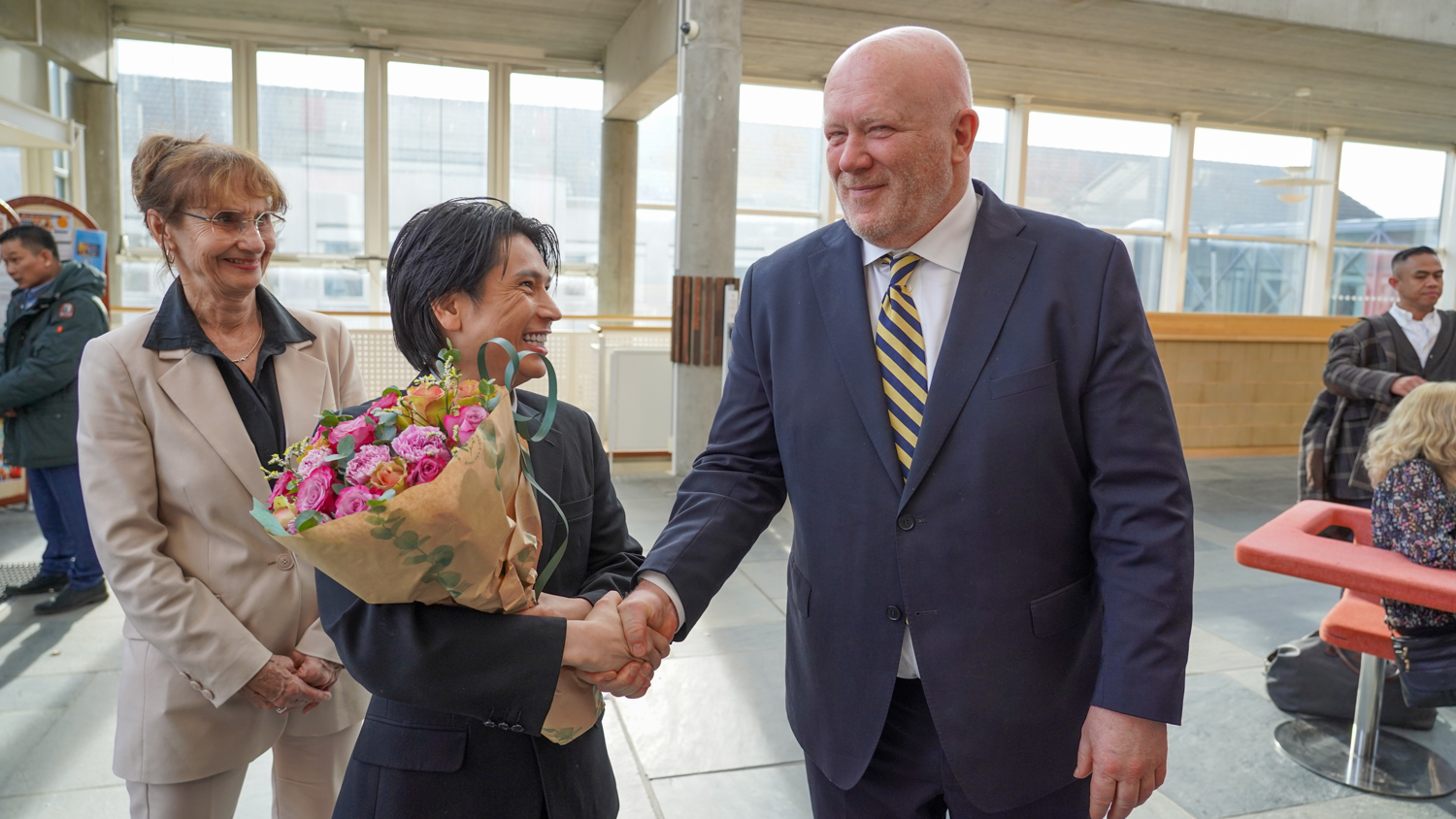 En mann tar en annen i hånda og gratulerer mens han holder en blomsterbukett og en dame står i bildet til venstre