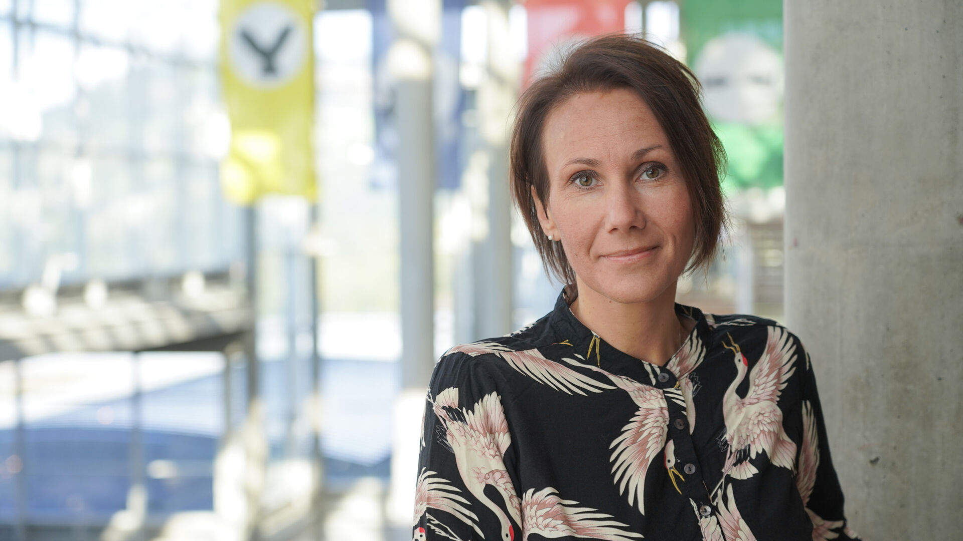 Portrettbilde av dame med mørkt, kort hår mot en vegg av store vinduer