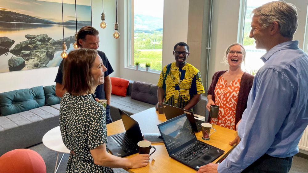 Bilde av kollegager som står og ler rundt et møtebord