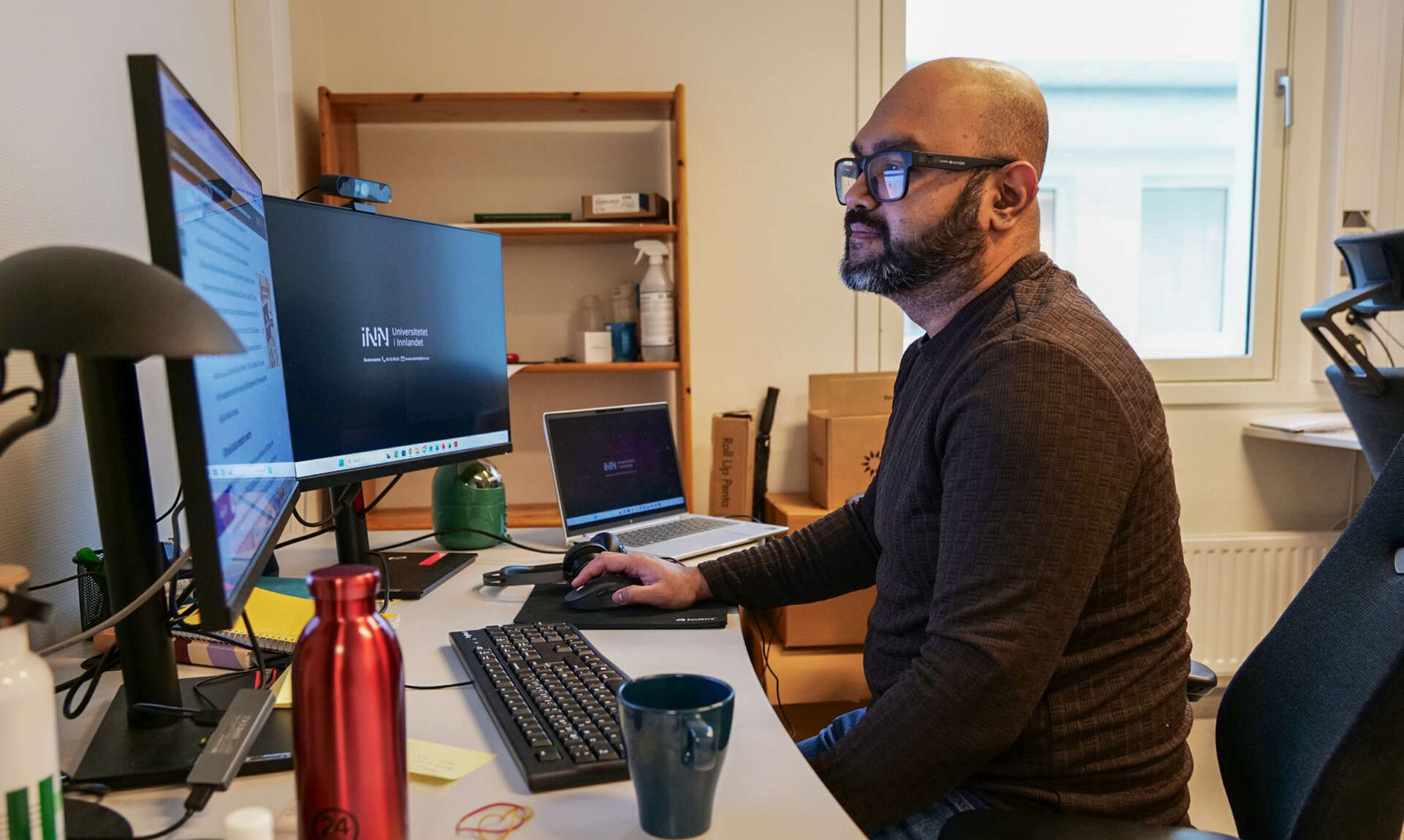 Mann med brun genser og briller sittet ved PCen sin ved et skrivebord