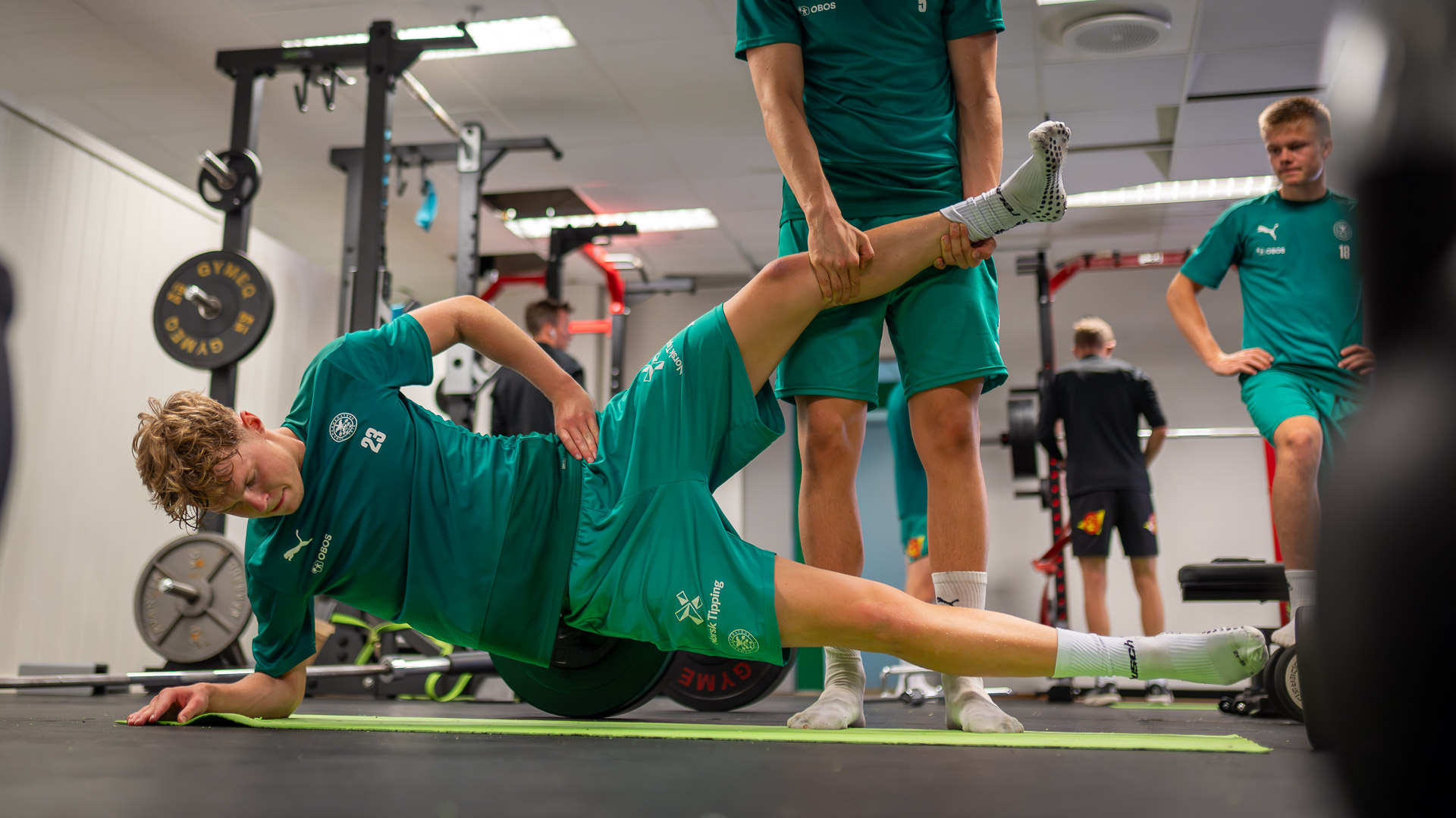 En fotballspiller ligger på siden på gulvet og får hjelp til å trene styrke.