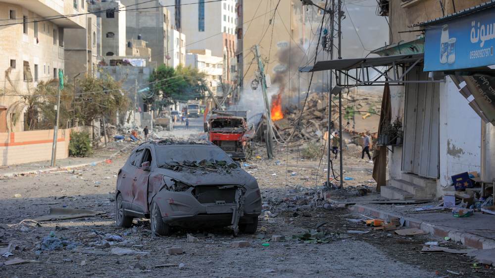 Bilde av utbrent bil foran ruiner og brann i Gaza