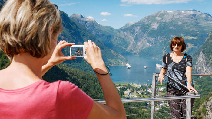 To damer står på et utsiktpunkt med Geiranger og fjorden i bakgrunnen