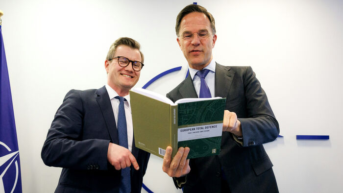 To menn står og leser i ei bok mot en hvot bakgrunn og NATO-emblemet