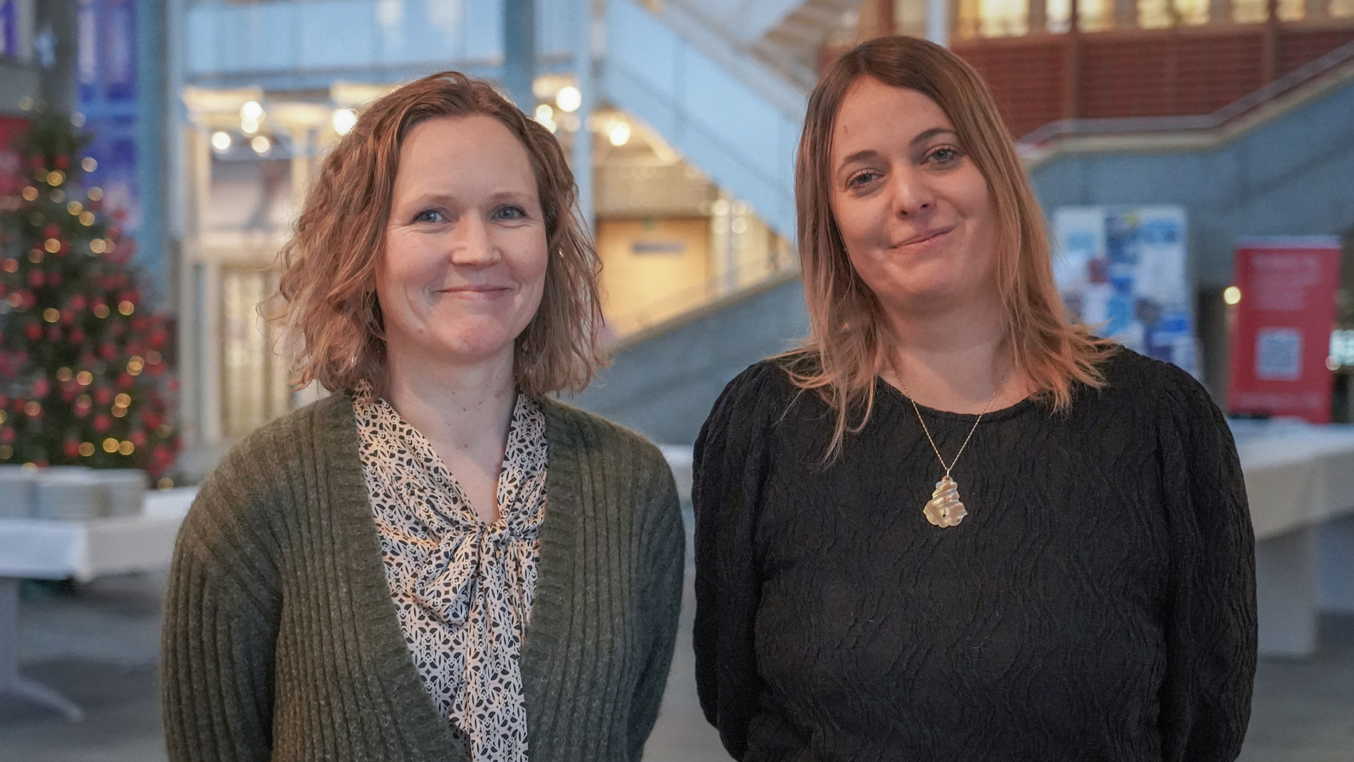 To damer ser i kamera i Glassgården på studiested Lillehammer på Universitetet i Innlandet med et juletre i bakgrunnen