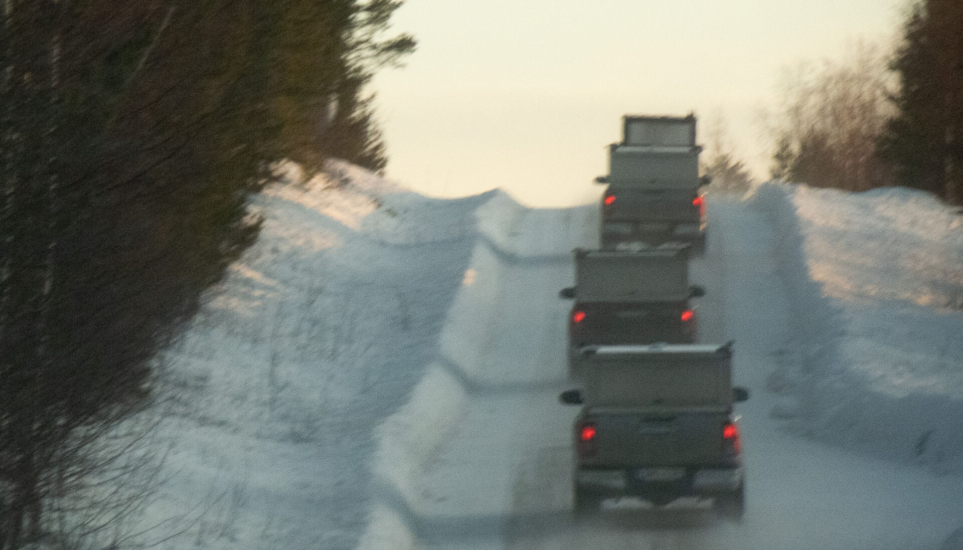 Four cars in a row pictured from the back driving on a snowy road with a forest to their left.