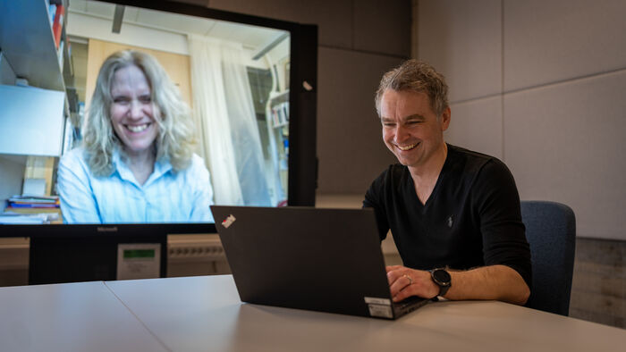 En person som sitter i et rom med PC foran en stor skjerm med en dame som ler