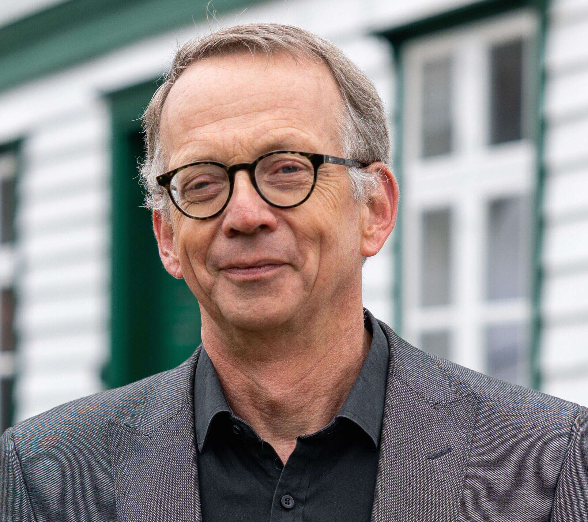 Eldre mann med briller, grå jakke og svart skjorte utenfor en bygning