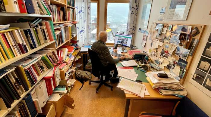 Professor sitter bak pulten sin på et kontor fullt av bøker og jobber 