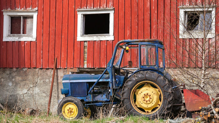 En sliten, gammel blå traktor står foran en rød låve med hvite vinduer