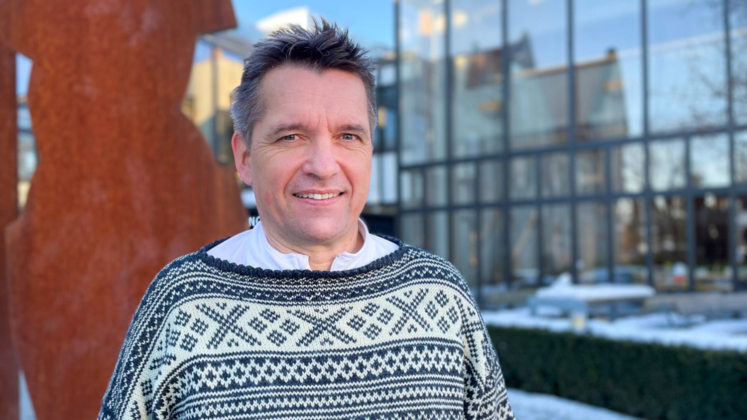 Jon Anders Græsli mot en rustfarget kunstskulptur og en vegg av store vinduer i bakgrunnen