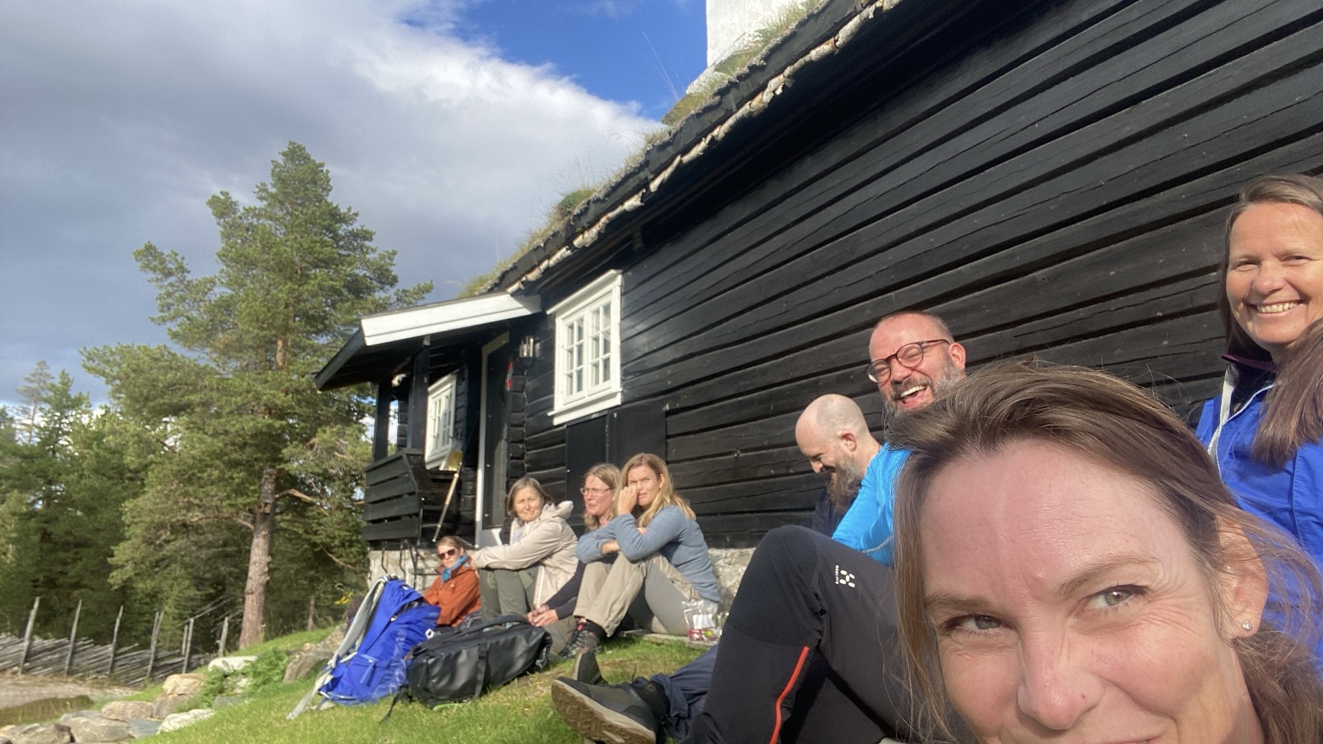 En gruppe mennesker sitter utenfor en hytte i sola. De smiler og er glade