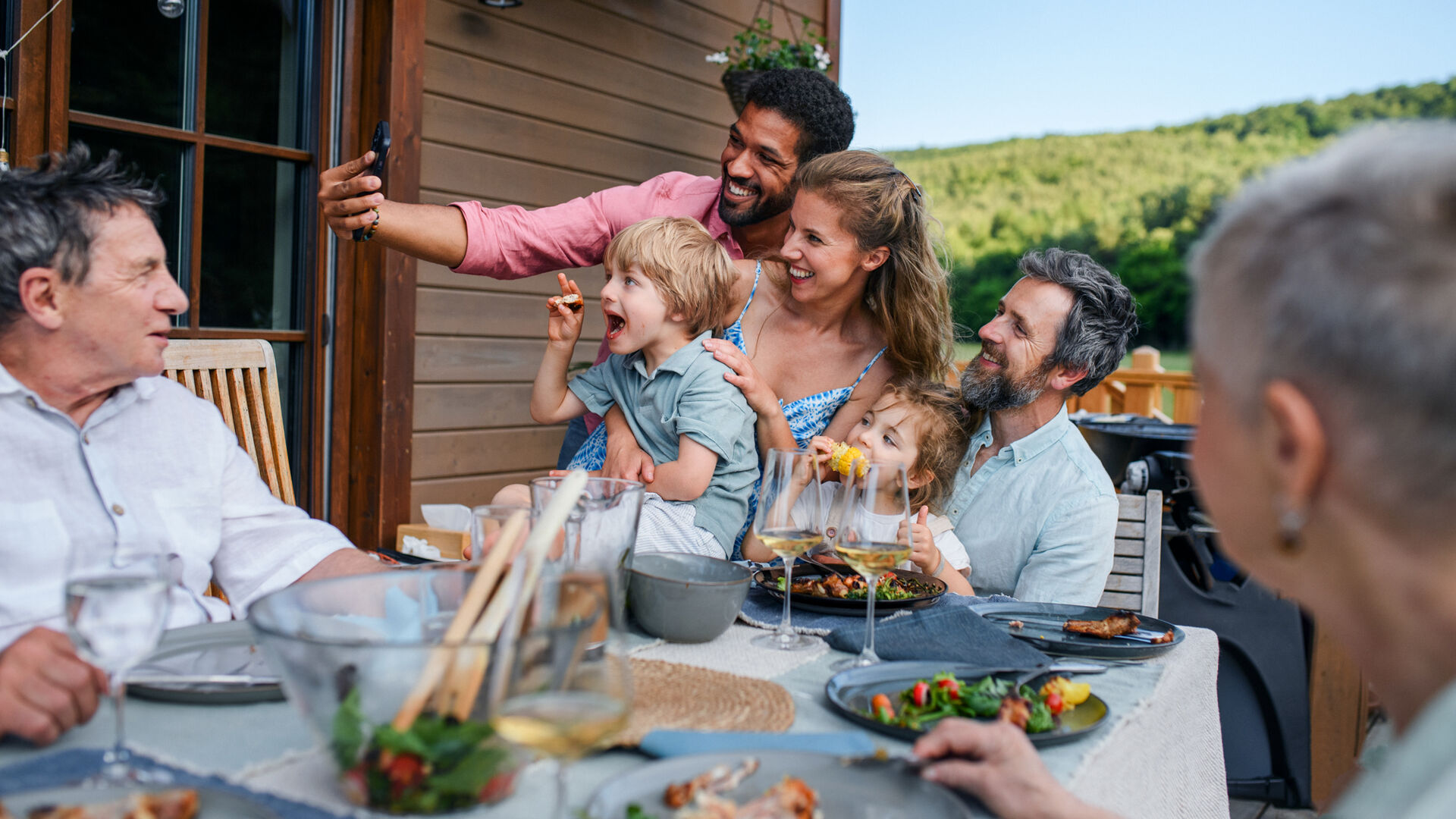 Flere mennesker i en familie sitter rundt et utebord på en veranda og noen tar en selfie