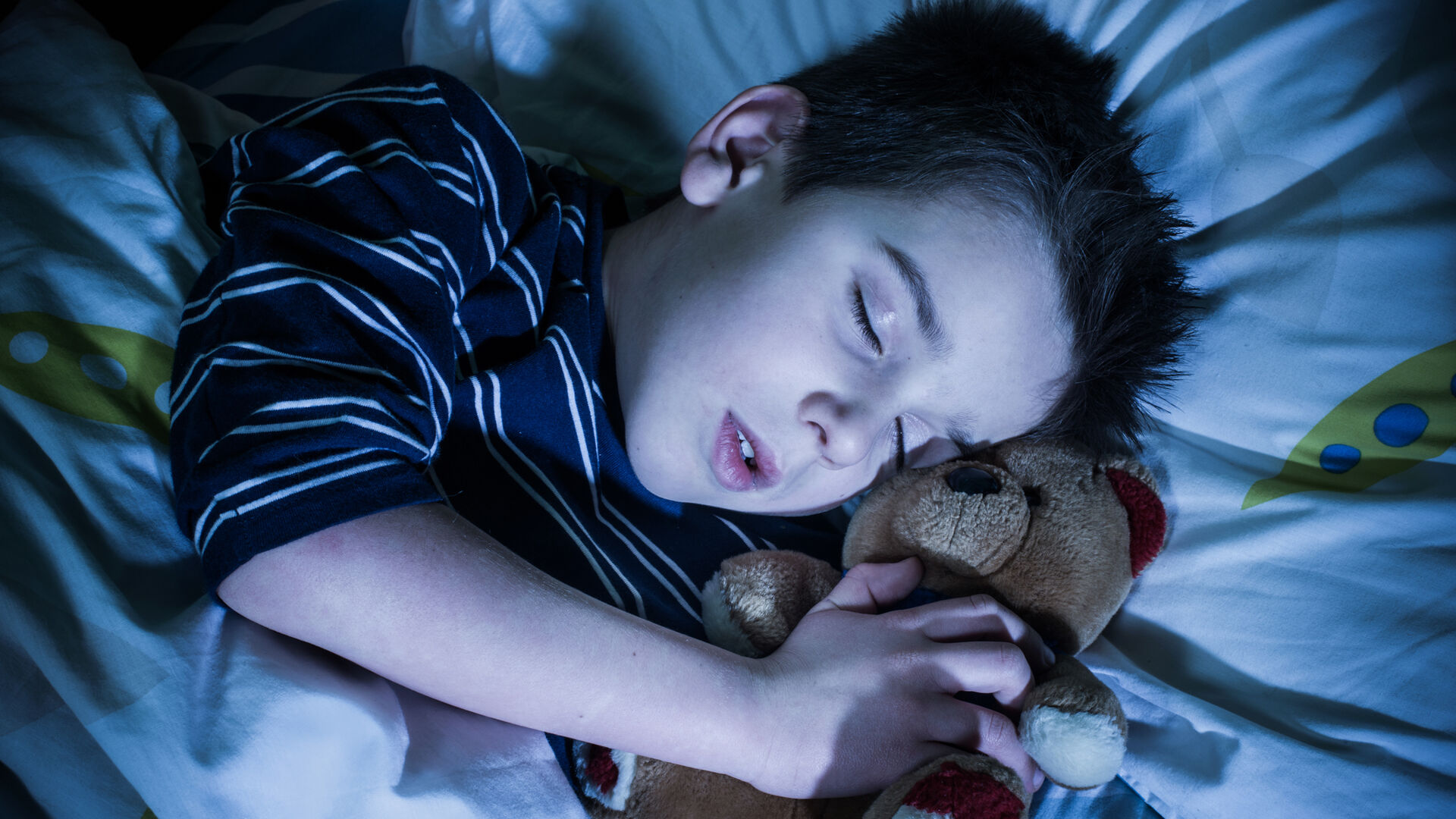 En liten gutt sover med bamse på armen i en seng