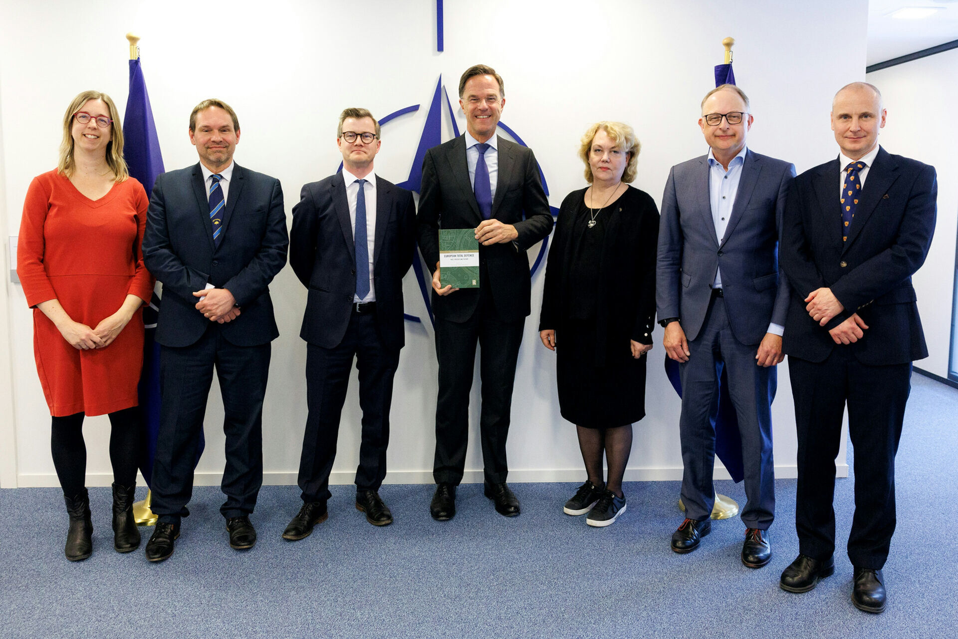 En gjeng forskere står sammen med NATO-sjefen foran NATO-flagg