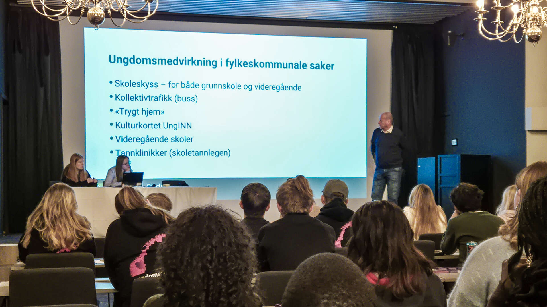 En mann og to ungdommer på en scene foran en stor skjerm