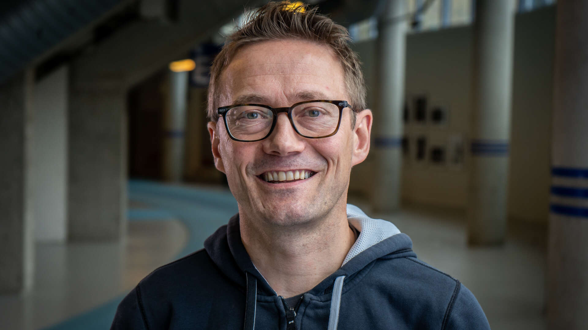 Mann med briller og hettejakke står inne på en innedørs løpebane i en gang