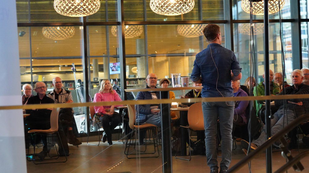 Foredragsholder sett bakfra. Publikum følger interessert med. 