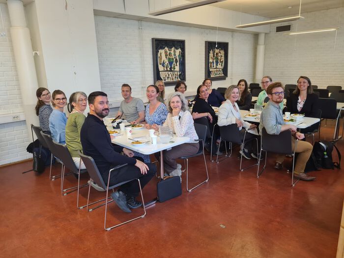 En kantine med to bord der det sitter personer på alle stolene som smiler til kamera
