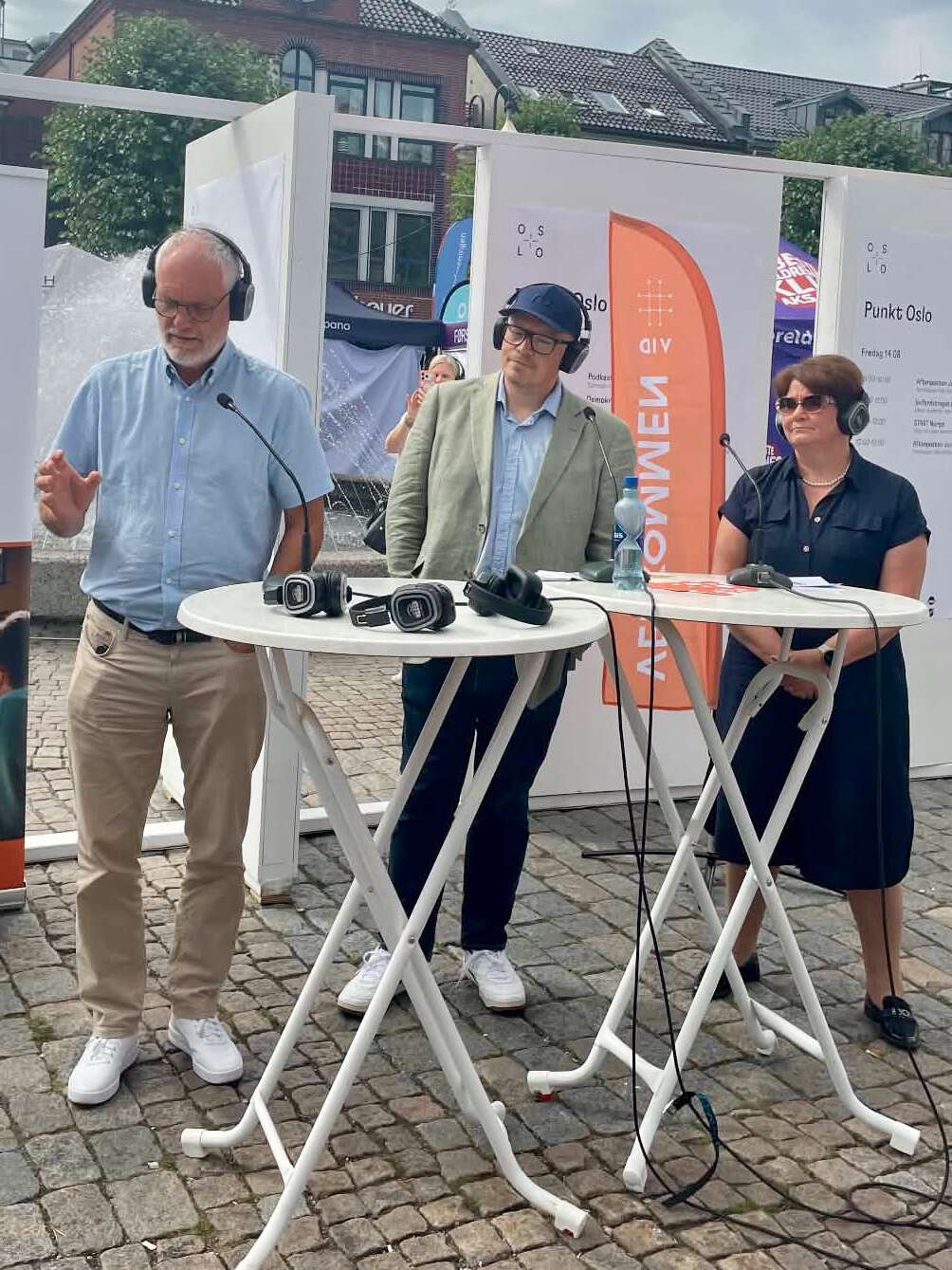 To menn og en dame står utendørs bak to bord og deltar i debatt. Alle har headset, og de deler på to mikrofoner. I bakgrunnen står et oransje beachflagg.