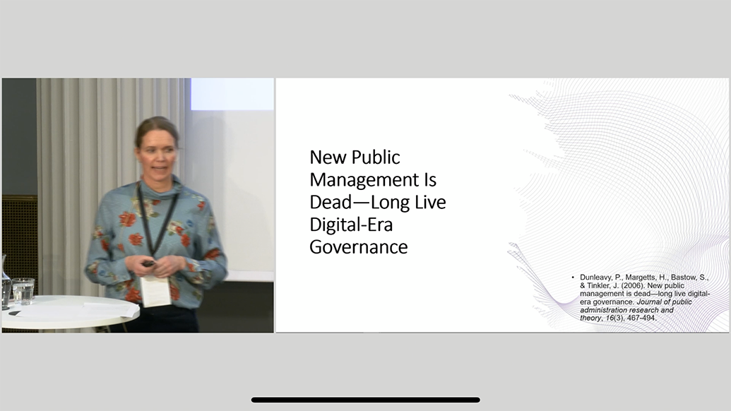 Dame i blomstrete genser bak et ståbord og ved siden av skjerm som viser powerpoint 