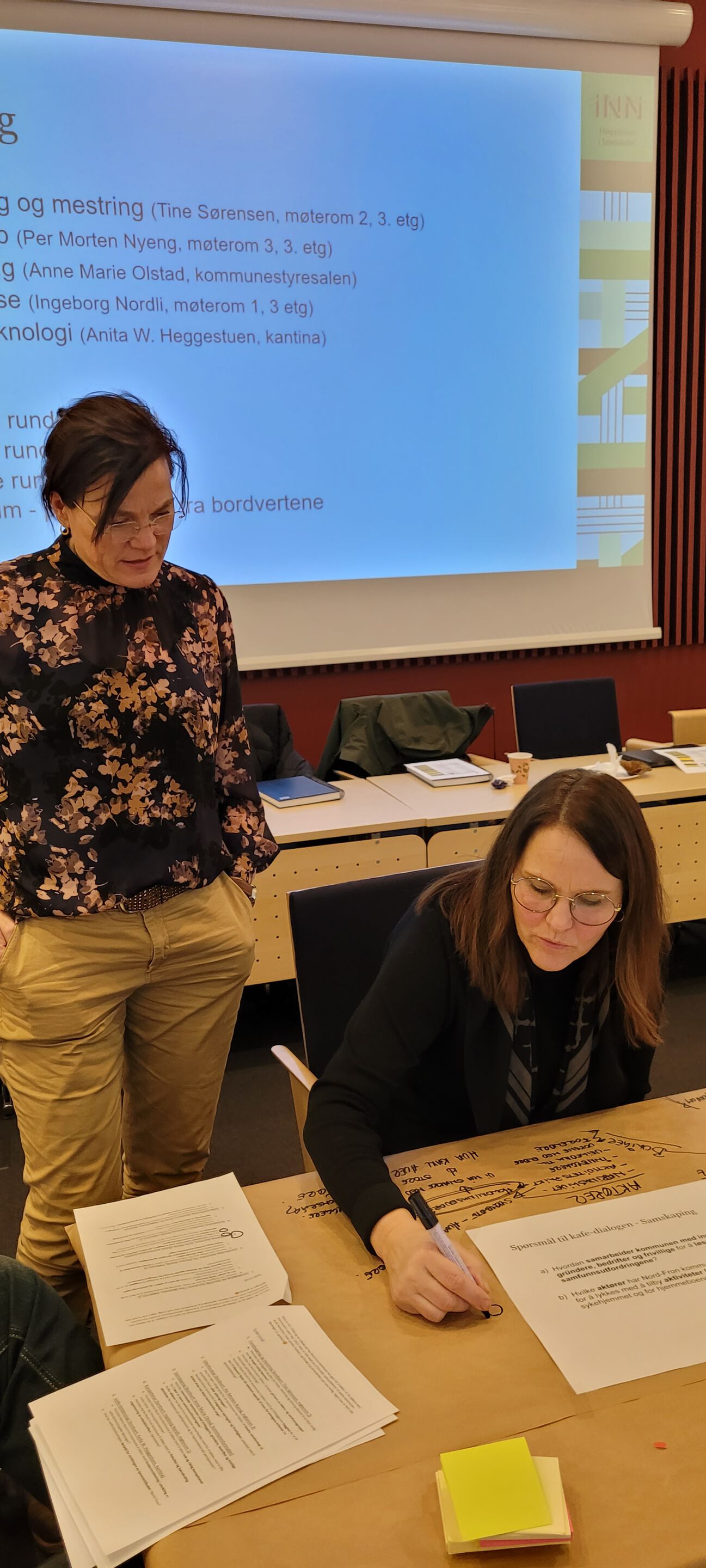Stående dame ser over skuldrene til sittende dame som skriver på en stor poster
