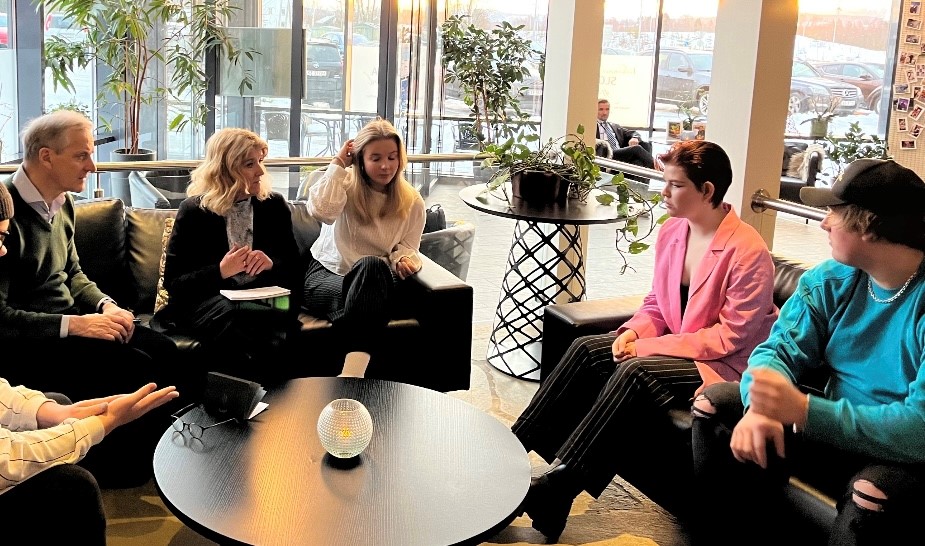 gruppe mennesker sittende på sofa foran store vinduer og med planter rundt. De ser ut som de er i en engasjerende diskusjon