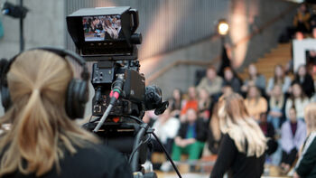 En jente med tv-kamera er fotografert bakfra mens hun filmer en gruppe personer som sitter i en trapp