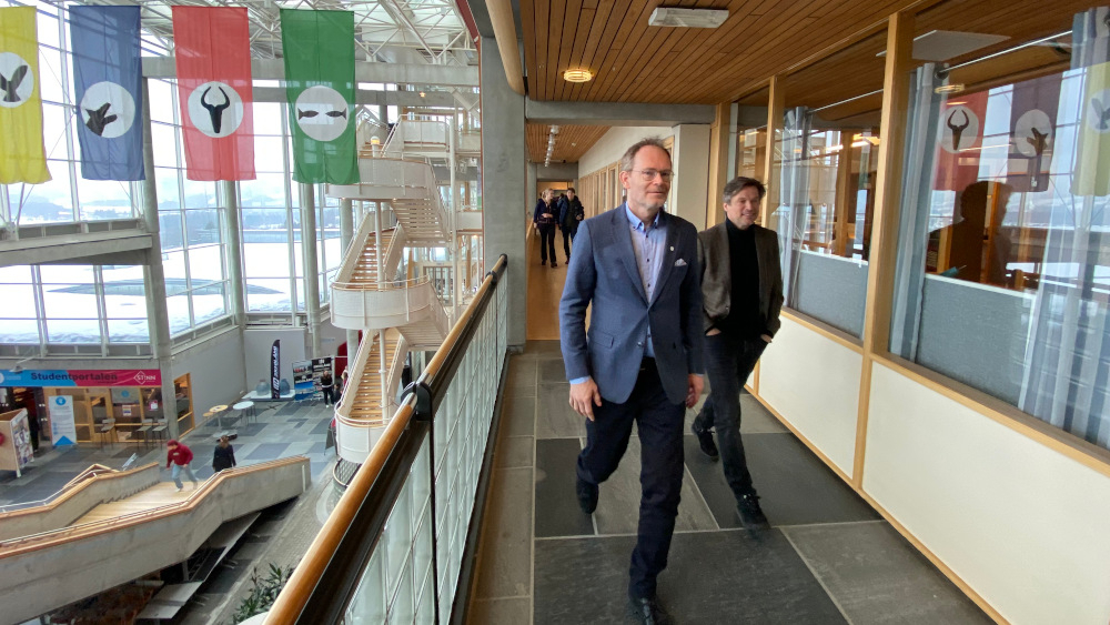 Oddmung Løkensgard Hoel og Leif Holst Jensen går i en korridor, i bakgrunne henger fire bannere i guilt blått rødt og grønt
