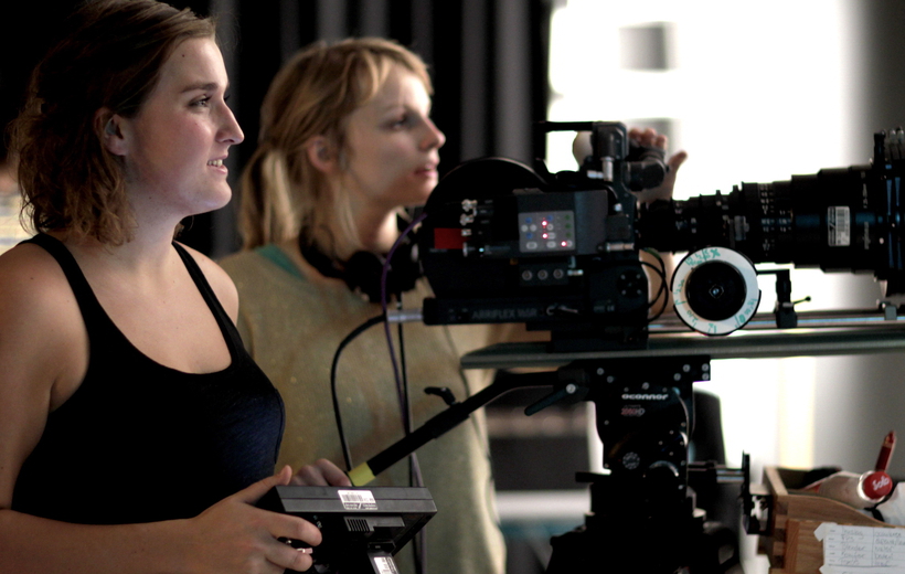 To jenter, regi og foto, står bak kamera i en opptakssituasjon.
