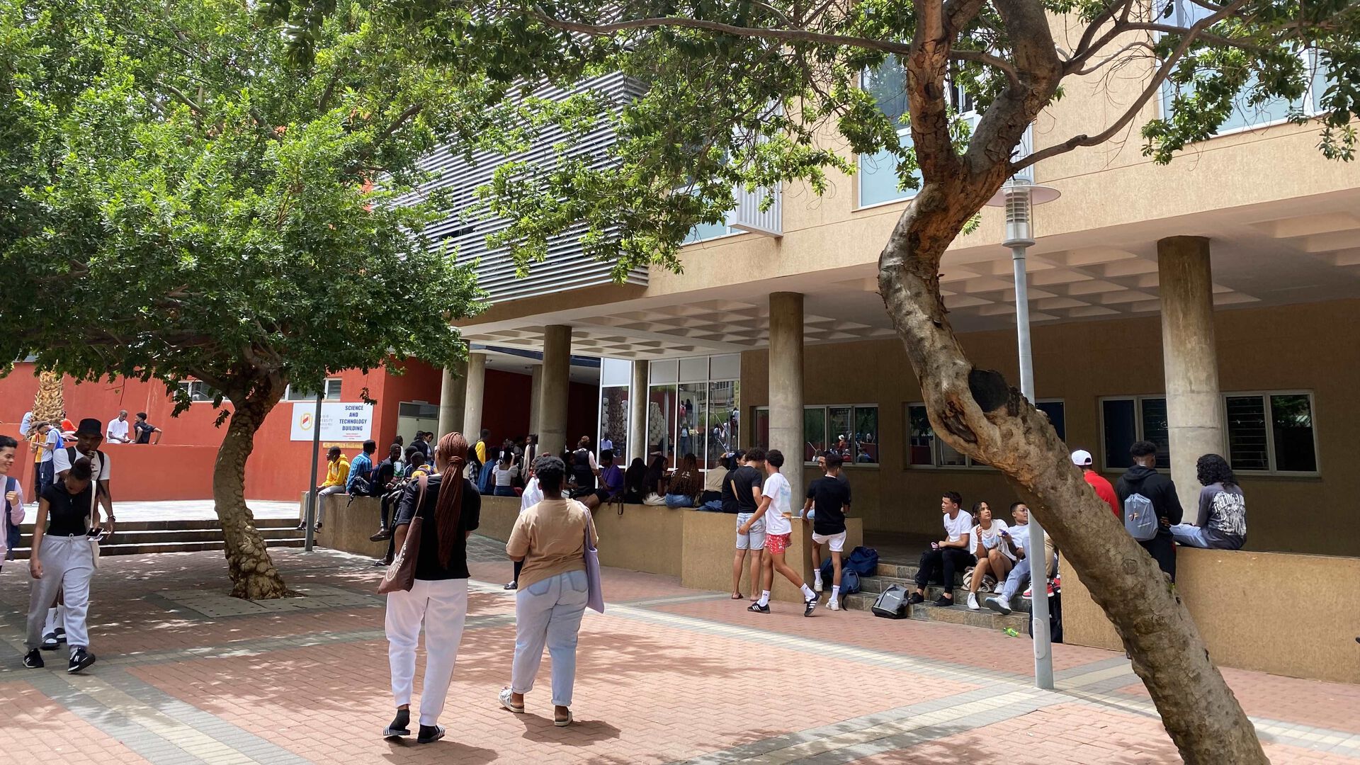 Yrende liv og mange mennesker som går forbi utendørs på campus i Namibia, med store trær som danner skygge.