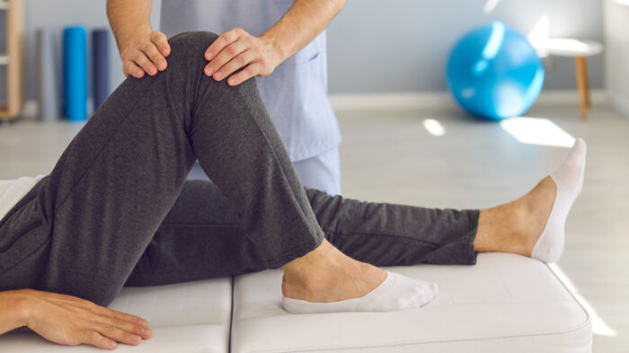 En fysioterapeut i blå klær holder hendene på kneet til en pasient som ligger på en benk