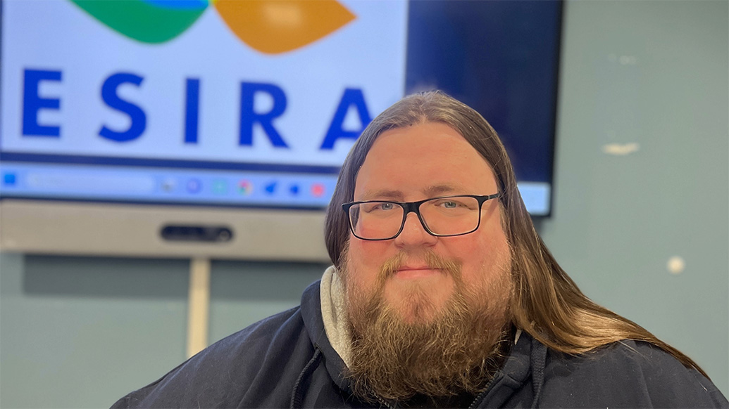 Mann med langt hår og sjegg smilende foran fargerik logo med bokstavene ESIRA