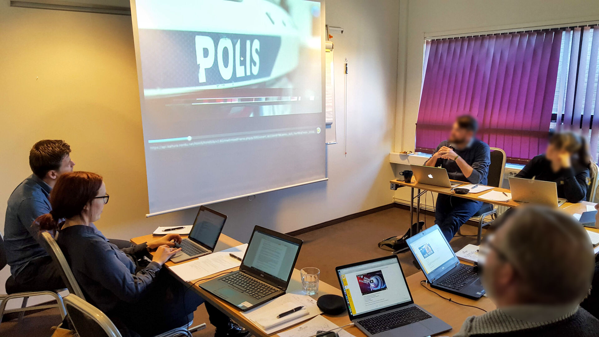 gruppe mennesker med hver sin pc ser på stor skjerm med bilde av svensk politibil