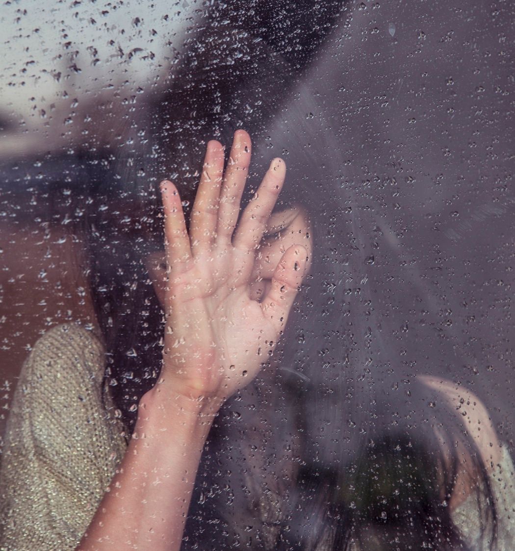 Jente med langt hår bak et vindu med regndråper, holder opp hånden sin mot vinduet.
