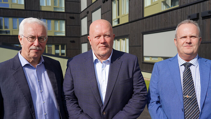 Tre personer i mørke dresser som står foran en ny bygning. 
