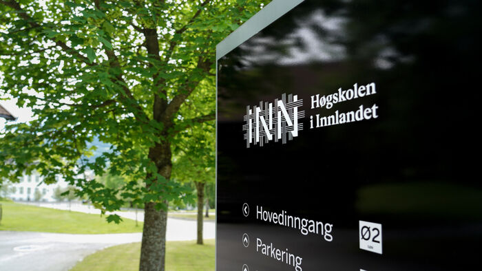 Et skilt med høgskolens logo utendørs, med et tre og en vei i bakgrunnen